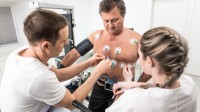 Eine medizinische Technologin und ein Technologe für Funktionsdiagnostik befestigen Messelektroden am Oberkörper eines Patienten.