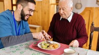 Ein Altenpfleger unterstützt einen Senior beim Essen an einem Tisch. Beide wirken entspannt und zufrieden.