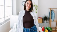 Eine Frau in einem weißen Kittel steht an einem Schreibtisch, auf dem eine Schüssel mit frischem Obst und Gemüse platziert ist.