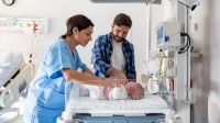 Eine Gesundheits- und Kinderkrankenpflegerin kontrolliert den Gesundheitszustand eines Neugeborenen. Daneben steht der Vater des Babys.