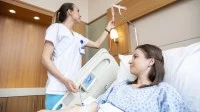 Eine Gesundheits- und Krankenpflegerin überprüft die Einstellung des Tropfes einer Patientin. Diese liegt im Krankenzimmer in ihrem Krankenbett.