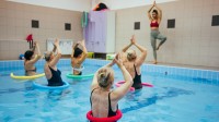 Eine Masseurin und medizinische Bademeisterin  leitet eine Gruppe von Frauen im Wasser an, die gemeinsam Yoga-Übungen im Schwimmbecken durchführen.