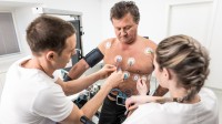 Eine medizinische Technologin und ein Technologe für Funktionsdiagnostik befestigen Messelektroden am nackten Oberkörper eines Patienten. 