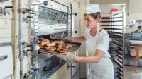 Eine Bäckerin holt frisch gebackene Brezeln und Brötchen aus dem Ofen.