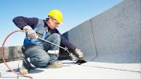 Ein Bauwerksabdichter bringt Dachpappe durch Erhitzen mit einem Gasbrenner auf einem Flachdach an. 