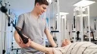 Ein Physiotherapeut unterstützt einen älteren Herrn in einem Fitnessraum dabei, seine Kraft in den Armen zu trainieren. 