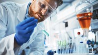 Ein chemisch-technischer Assistent untersucht in Schutzkleidung im Labor eine Probe mit einer hellblauen Reagenz. Weitere Reagenzien stehen auf dem Tisch.