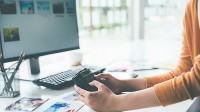 Hände halten eine Kamera vor einem Computerbildschirm. Auf dem Tisch befinden sich Fotos, eine Tastatur und ein Stiftehalter.