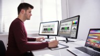 Ein Kommunikationsdesigner arbeitet mit einem Zeichenpad, zwei Bildschirmen und einem Laptop an einer neuen Werbung. 
