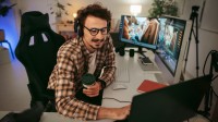 Ein Mediendesigner arbeitet in seinem Büro. Er hält einen To-go-Becher in der Hand. Auf dem Schreibtisch befinden sich zwei Bildschirme, ein Laptop und weitere Technik.