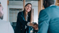 Eine Frau im Businessoutfit begrüßt einen ausländischen Mann per Handschlag. Ein weiterer Mann beobachtet das Geschehen.