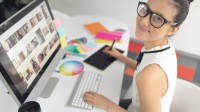 Eine glückliche gestaltungstechnische Assistentin arbeitet an ihrem Computer. Auf dem Bildschirm werden verschiedene Fotos angezeigt, zudem liegen Farbtafeln auf ihrem Schreibtisch. 