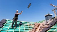 Ein Dachdecker wirft seinem Kollegen auf dem Dach einen Ziegel zu.