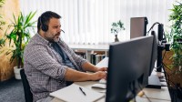 Ein übergewichtiger Mann mit einem Headset auf dem Kopf sitzt im Büro an seinem Schreibtisch und tippt etwas in die Computertastatur. 