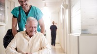 Ein glücklicher Gesundheits- und Krankenpflegehelfer fährt einen älteren Mann im Rollstuhl über den Flur und trägt dabei die Tasche des Mannes.