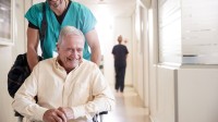 Ein Gesundheits- und Krankenpflegehelfer bringt einen entlassenen Patienten mit dem Rollstuhl zum Ausgang des Krankenhaus.