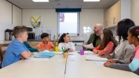 Ein Sozialassistent kümmert sich in einer Gemeinschaftsküche um eine Gruppe Kinder verschiedener ethnischer Herkunft. 