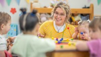 Eine lächelnde sozialpädagogische Assistentin / Kinderpflegerin spielt mit vier Kleinkindern am Tisch in einem Kinderhort.