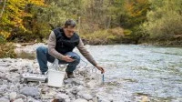 Ein umweltschutztechnischer Assistent hockt neben einem Flusslauf und hält ein Messgerät in das Wasser.