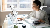 Eine kaufmännische Assistentin / Wirtschaftsassistentin für Fremdsprachen sitzt an ihrem Schreibtisch im Büro und tippt Zahlen, die sie mit Hilfe eines Taschenrechners berechnet, in ihren Laptop. 