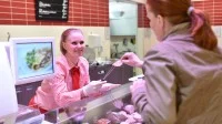 Eine Fachverkäuferin im Lebensmittelhandwerk bietet einer Kundin an der Fleischtheke ein Stück Wurst zum Probieren an.