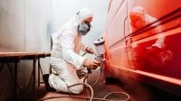 Ein Fahrzeuglackierer in Schutzausrüstung besprüht ein Auto mit rotem Lack.