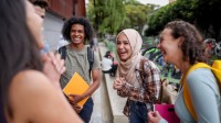 Fünf junge Menschen mit unterschiedlicher ethnischer Herkunft stehen im Kreis und lachen. Sie halten Schulsachen in den Händen.