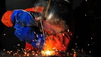 Ein Schweißer in orangener Schutzkleidung, blauen Handschuhen und Helm, der etwas schweißt. Der Hintergrund ist schwarz und Funken sprühen.