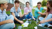Eine Gruppe von fünf jungen Leuten sitzt bei sommerlichen Temperaturen auf einem Grünstreifen vor einem Gebäude. Die Personen haben ihre Notebooks, Schreibunterlagen sowie Getränke und Snacks dabei.