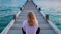 Eine Frau sitzt mit dem Rücken zum Betrachter auf einem Bootssteg und blickt zum Wasser sowie Horizont.