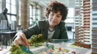 Eine Frau mittleren Alters sitzt in einem Büro vor einem Modell und berührt dieses mit der rechten Hand. Auf dem Modell ist ein Wohnblock, eine Freizeitanlage, eine Grünfläche und eine Straße gebaut.