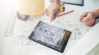 Auf einem Tisch liegt eine Bauzeichnung, auf der wiederum ein Tablet mit einem Hologramm eines Hauses sowie ein Lineal, Helm und Stift liegen. Eine Hand berührt das Hologramm.