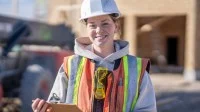 Eine junge Frau mit Bauhelm und Warnweste steht auf einer Baustelle und hat ein Klemmbrett in der Hand.