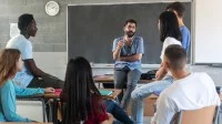 Ein Berufsschullehrer lehnt am Pult und diskutiert mit einigen Schülerinnen und Schülern.