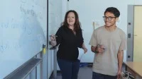 Eine Lehrerin steht mit einem Auszubildenden an der Tafel und erklärt eine Aufgabe. 