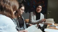 Drei Schülerinnen sitzen an einem Tisch mit Büchern und Schreibmaterial.