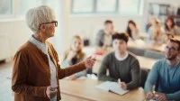 Eine alte Lehrerin steht im Klassenzimmer vor einer Gruppe von Schülerinnen und Schülern und erklärt den Unterricht. Die Lernenden hören aufmerksam zu und beteiligen sich am Gespräch.