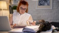 Eine Lehrerin arbeitet zuhause am Laptop und macht sich Notizen in ein Heft. Auf dem Schreibtisch liegt eine Katze.
