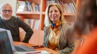Drei Personen sitzen in einem Büro. Auf dem Tisch sind Schreibutensilien, ein Laptop und ein Handy.