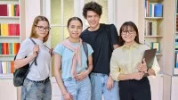 Zwei Schülerinnen, ein Schüler und eine Lehrerin stehen in der Bibliothek und lächeln in die Kamera.