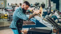 Ein Medientechnologe Siebdruck bedient eine Siebdruckmaschine zum Bedrucken von T-Shirts.