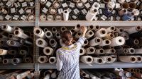 Eine Produktgestalterin - Textil wählt Stoff aus einem Stapel von Rollen in einer Textilwerkstatt aus.