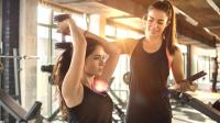Eine Sportfachfrau leitet im Fitnessstudio eine Kundin beim Hanteltraining an.