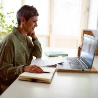 Eine Frau sitzt vor einem Laptop und lächelt, neben ihr ein Notizheft.