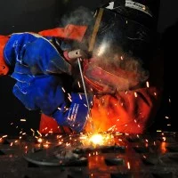 Ein Schweißer in orangener Schutzkleidung, blauen Handschuhen und Helm, der etwas schweißt. Der Hintergrund ist schwarz und Funken sprühen.