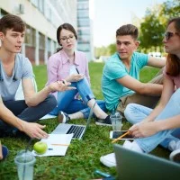 Eine Gruppe von fünf jungen Leuten sitzt bei sommerlichen Temperaturen auf einem Grünstreifen vor einem Gebäude. Die Personen haben ihre Notebooks, Schreibunterlagen sowie Getränke und Snacks dabei. 