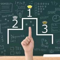 Ein Siegertreppchen mit den Plätzen1 bis 3, eine Kinderhand zeigt darauf. Im Hintergrund eine Schultafel mit Zeichnungen und Beschriftungen.