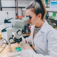 Eine junge Auszubildende sitzt an einem Mikroskop und arbeitet an einer Leiterplatte.