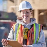 Eine junge Frau mit Bauhelm und Warnweste steht auf einer Baustelle und hat ein Klemmbrett in der Hand.
