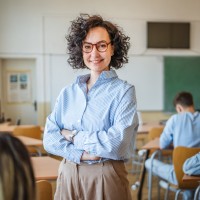 Eine Lehrerin steht mit verschränkten Armen in ihrer Klasse. Sie wirkt ruhig, gelassen und kompetent.